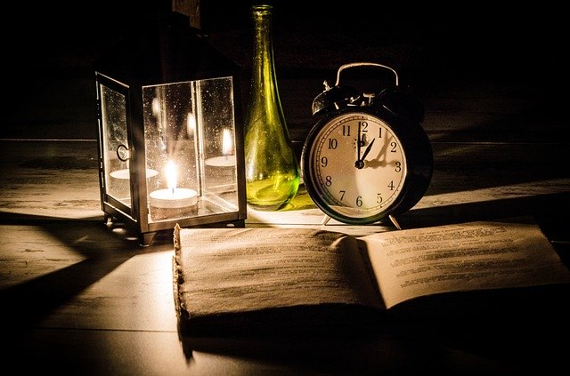 A lit candle on a desk next to a clock and an open book